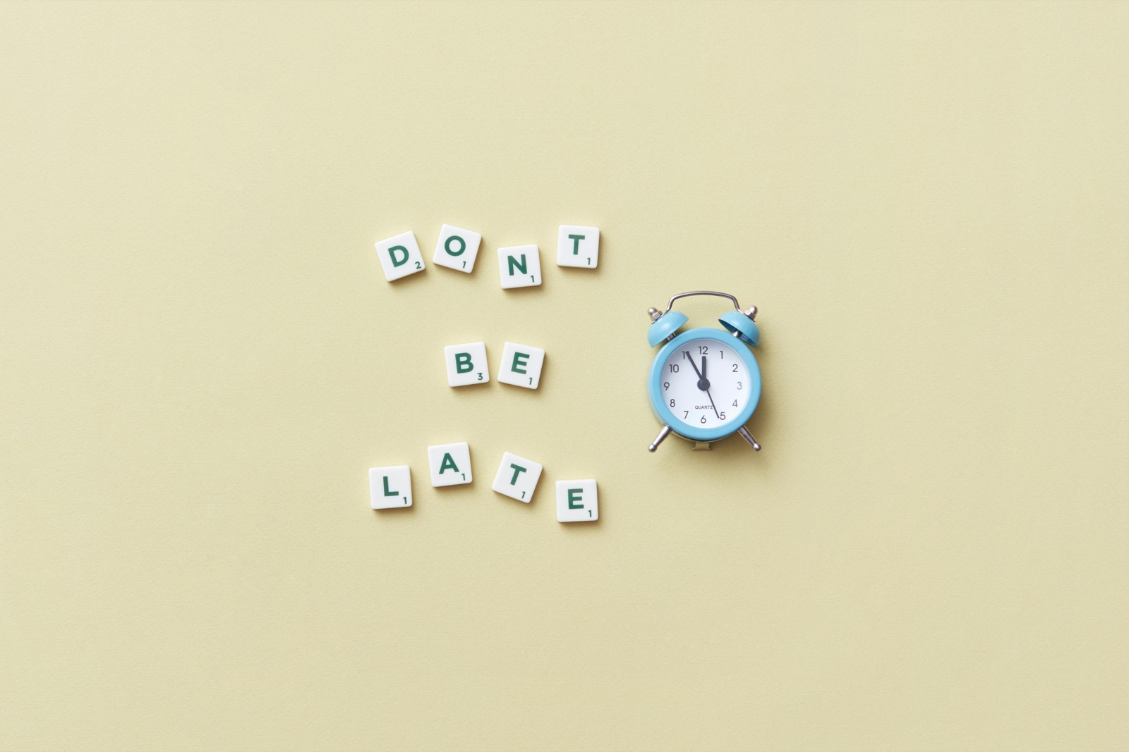 Scrabble tiles spelling DON'T BE LATE next to a small blue alarm clock on a yellow background